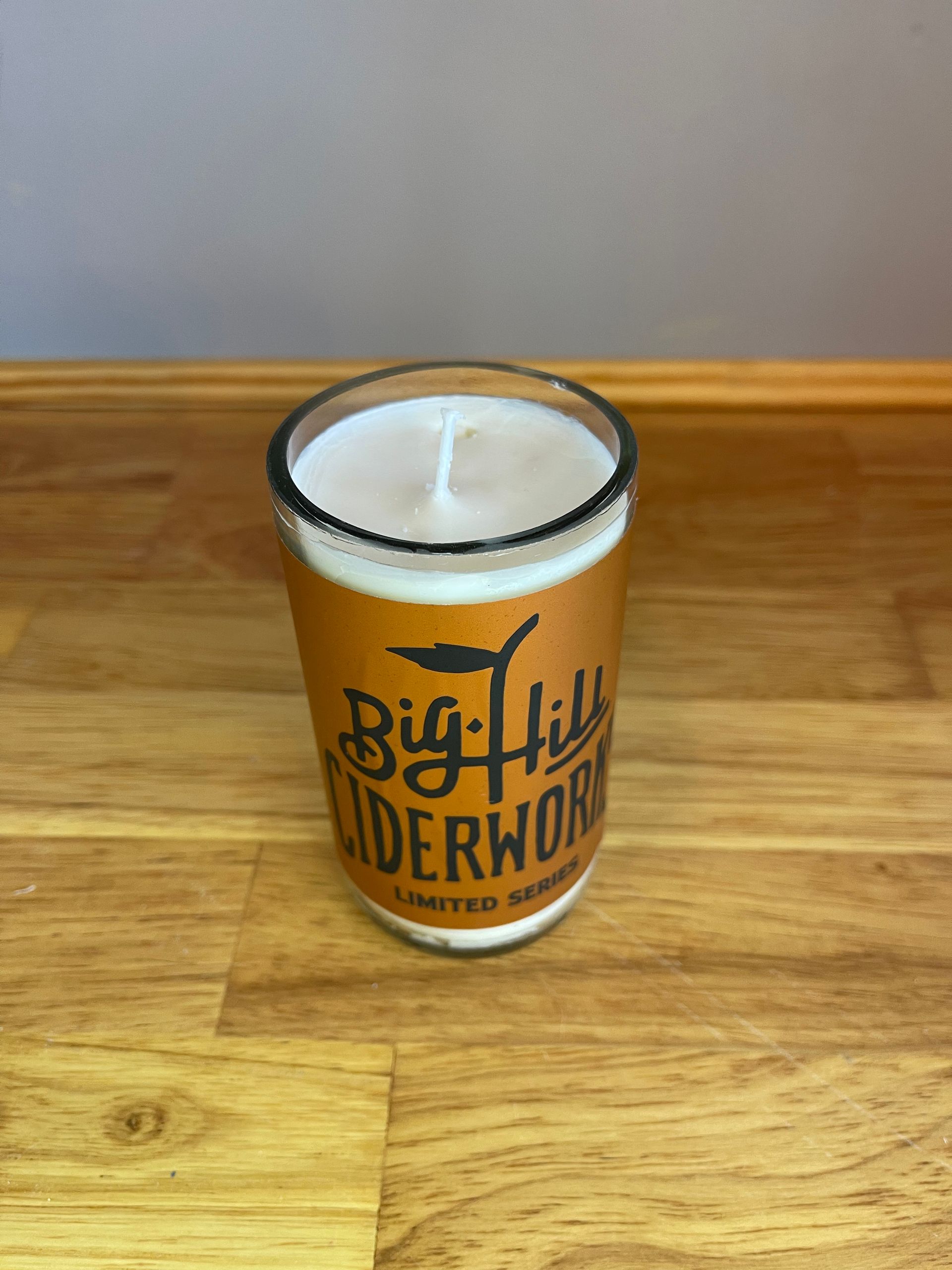 White candle in a clear glass jar with orange Big Hill Ciderworks label on a wooden surface.