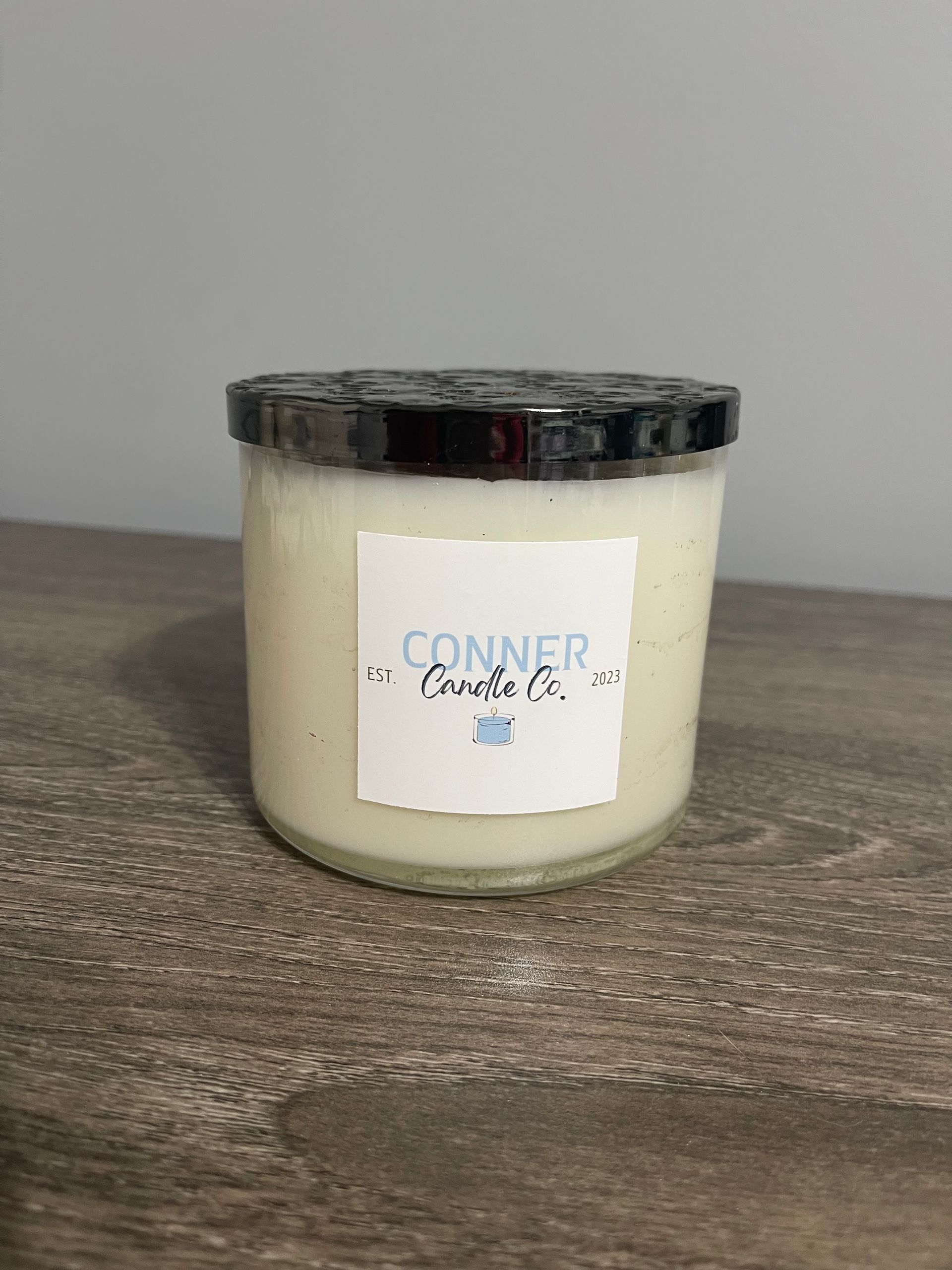 Cream-colored candle in a glass jar with a black lid and white label that says "Conner Candle Co." on a wooden table.
