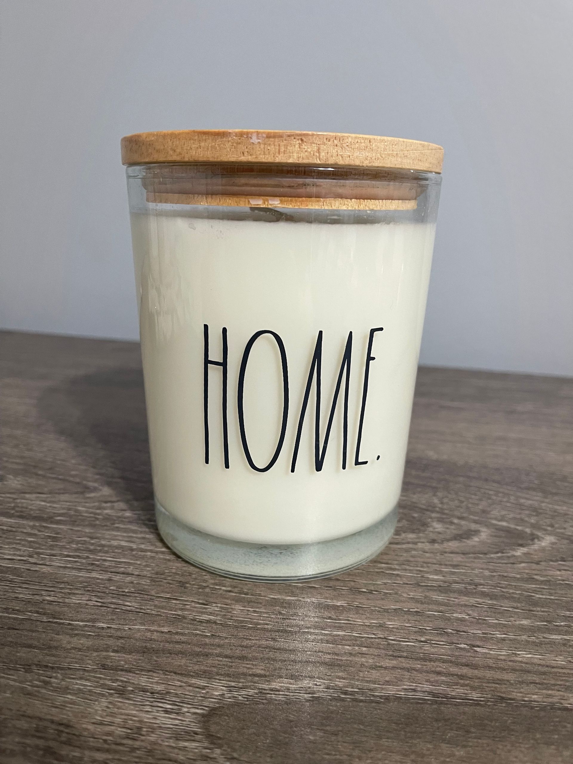 White candle in a glass jar with a wooden lid; the word "HOME" is printed on the jar.
