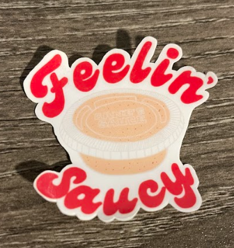 Sticker with red "Feelin' Saucy" text around a white container of sauce, on a gray surface.