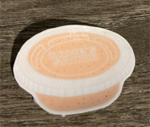 A single-serve cup of sauce with the text "KINDER'S SAUCE" visible inside. The sauce is peach-colored.