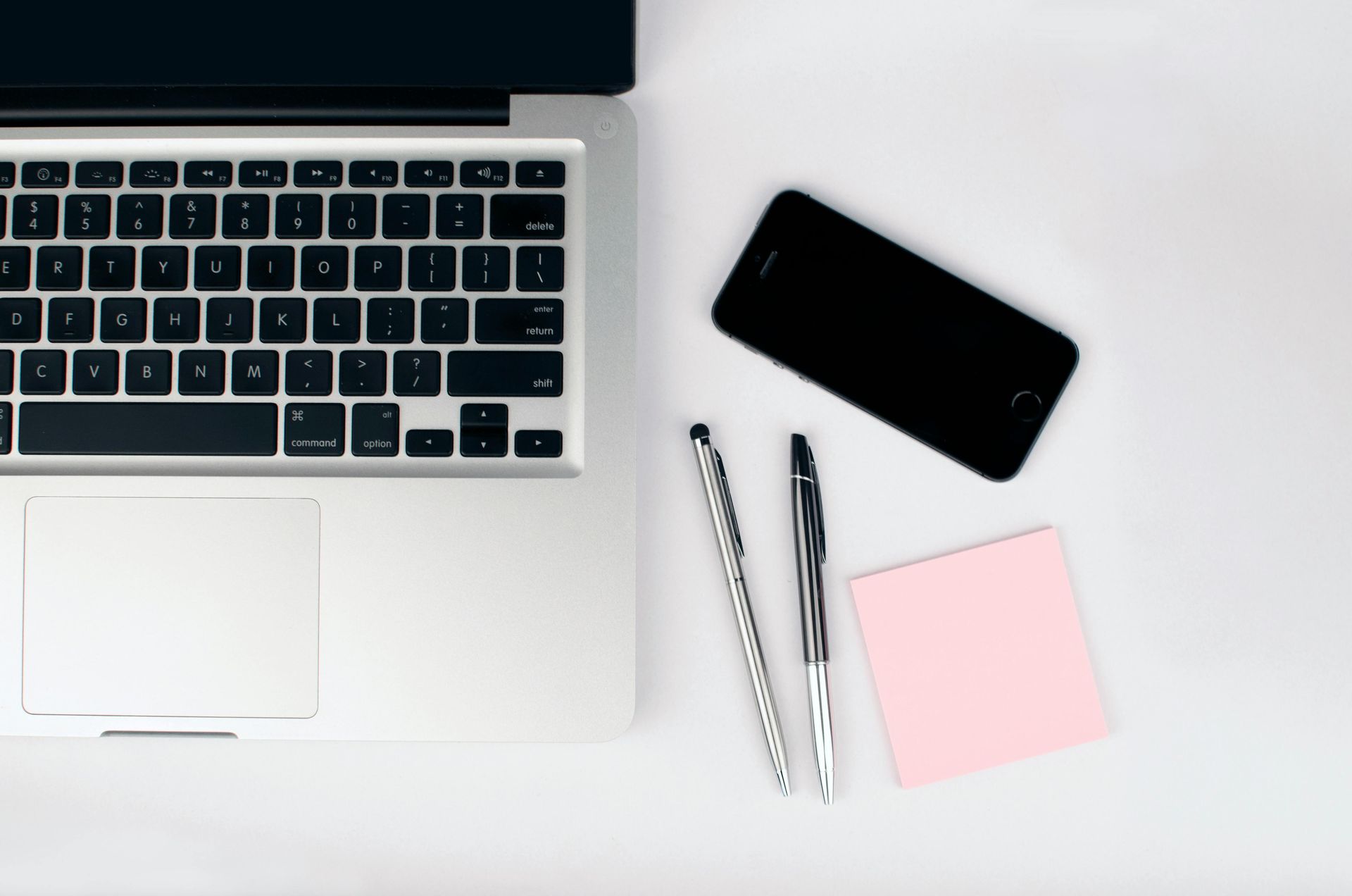 Laptop, smartphone, pens, and sticky note on a white surface.