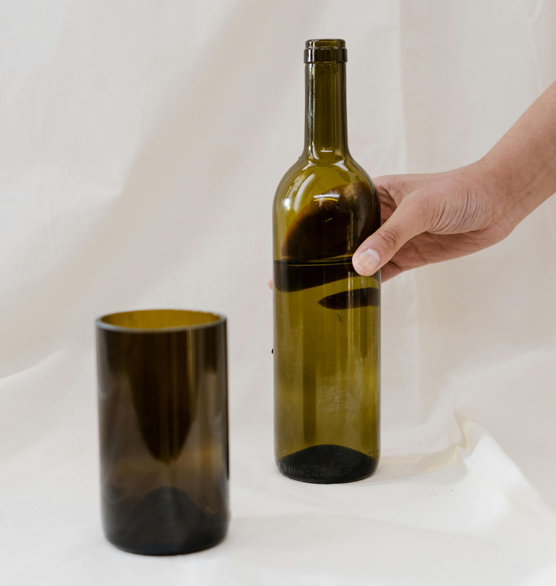 Hand holding an olive-green wine bottle beside a dark amber glass on a white background