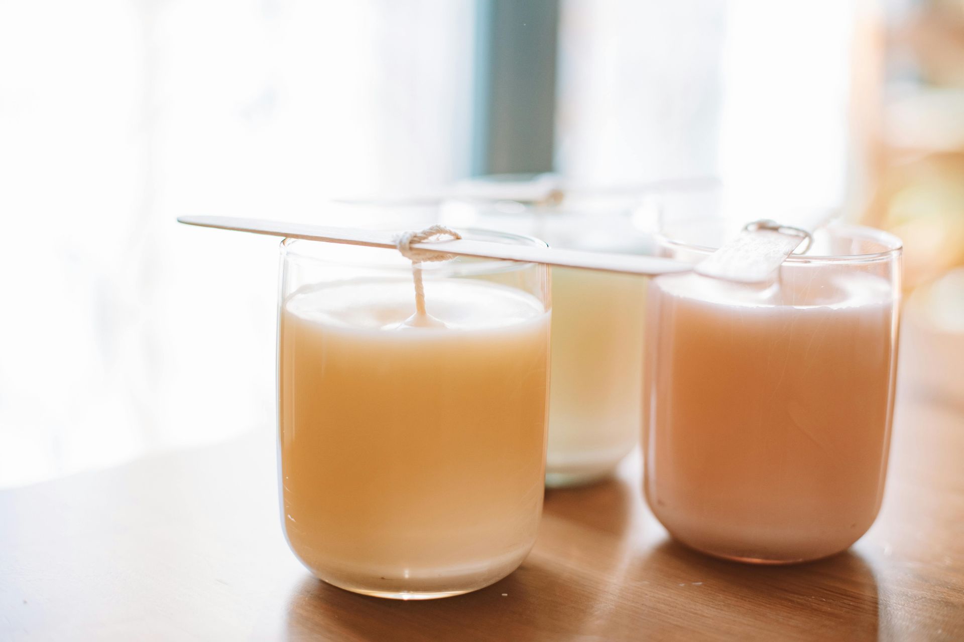 Three candles in glass jars, various pastel colors, wooden sticks holding wicks, on a wooden surface.