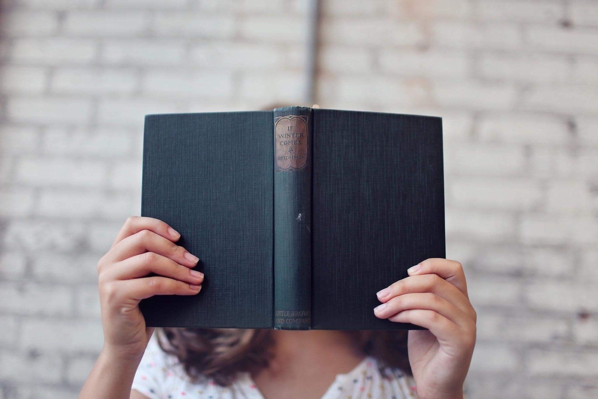 Person holding open dark green book in front of face against a white brick wall.