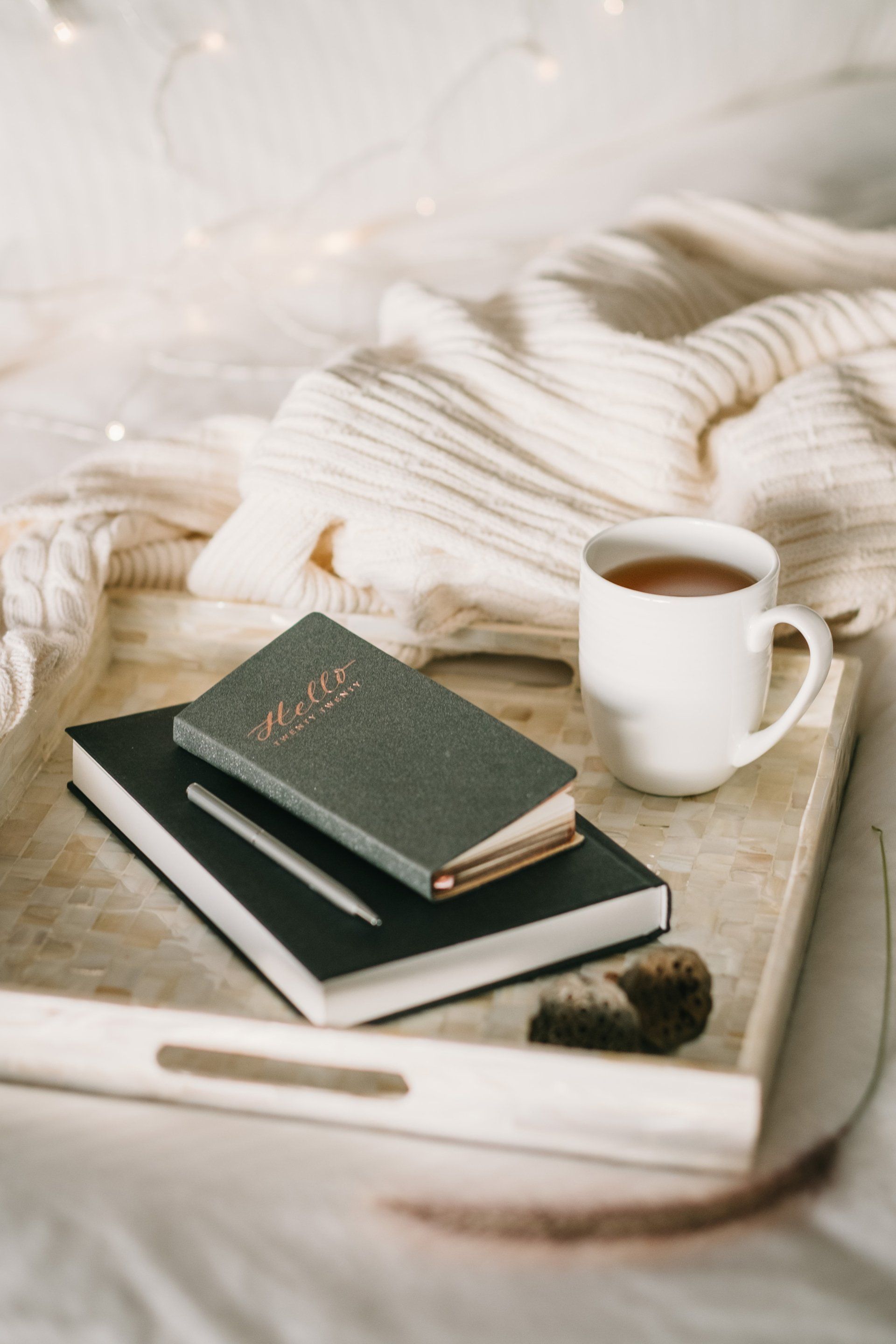 Tray with mug of tea, books, pen, and knitted blanket. White bedding and cozy setting.