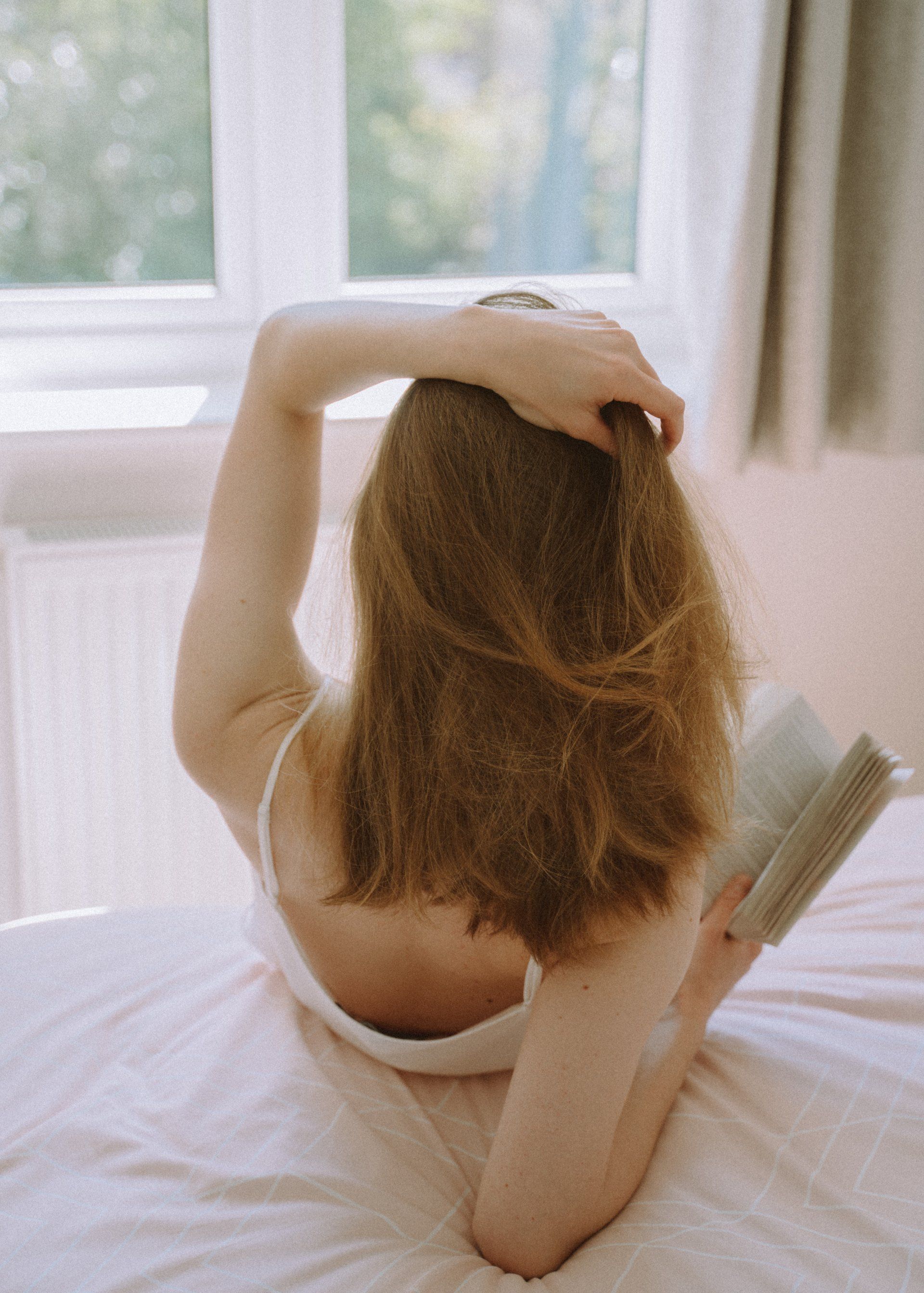Woman with long brown hair, wearing a white tank top, sitting on a bed reading a book, near a window.