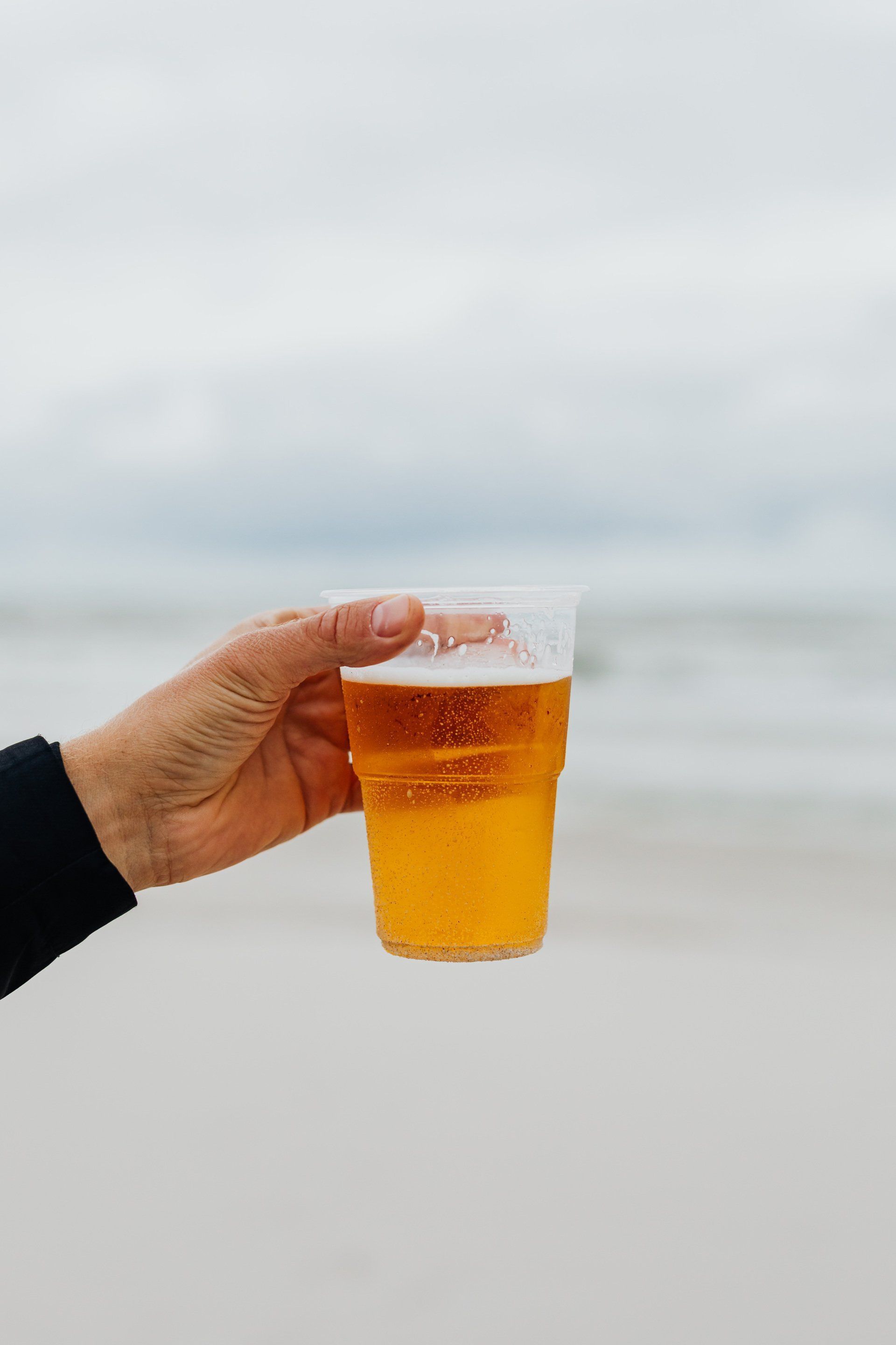 Hand holding a plastic cup of beer on a beach with the ocean in the background.