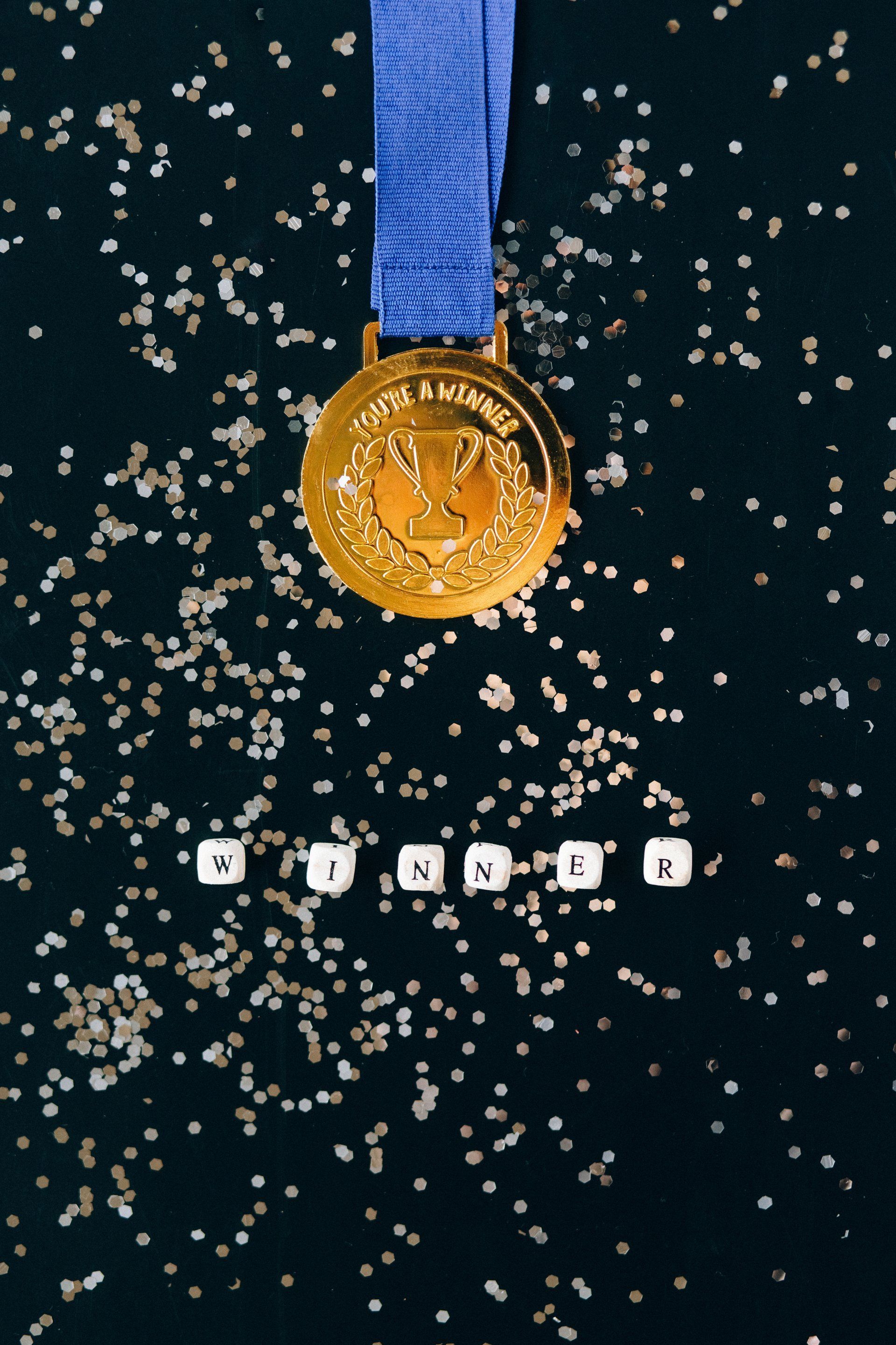 Gold medal with blue ribbon above, white letter blocks below spell
