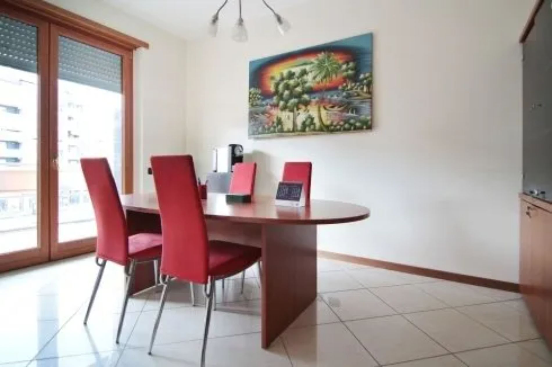 Sala da pranzo con tavolo in legno, sedie rosse e un quadro alla parete. La stanza ha una grande finestra.