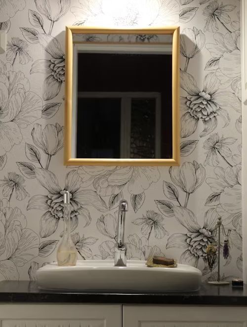 Bathroom with floral wallpaper, a square mirror with a wooden frame, and a white sink.
