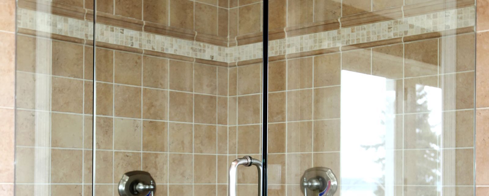 A glass shower door with a metal handle, set against a tiled wall featuring a decorative horizontal border strip.
