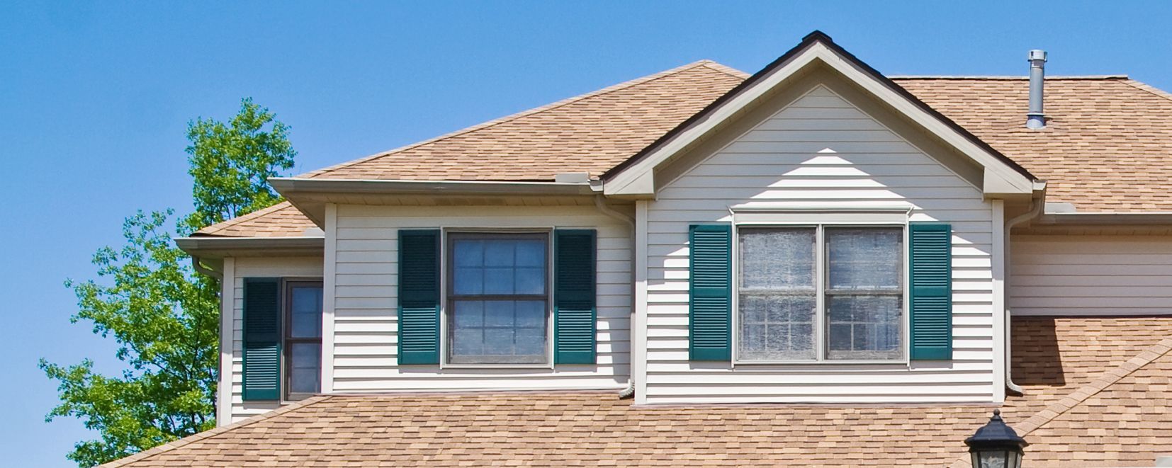 A beige two-story house with a brown shingled roof, a front-facing gable, and windows with green shutters under a blue sky.