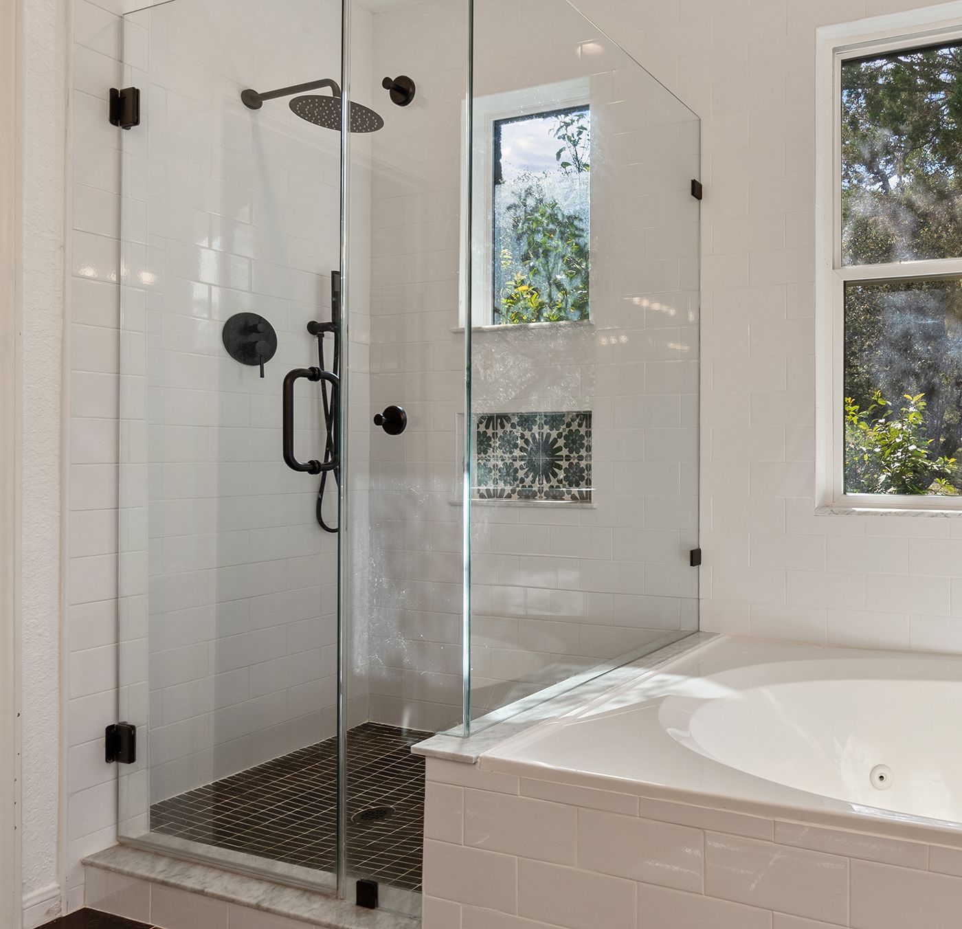 Glass shower enclosure with black fixtures and tile floor. Adjacent bathtub and window in a white bathroom.