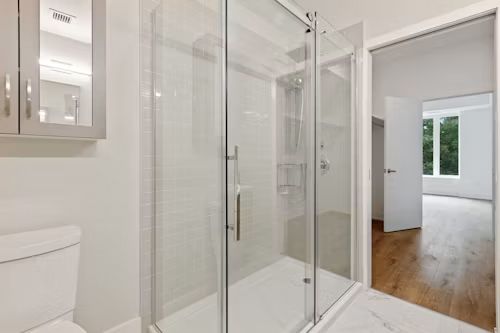 Bright bathroom with glass shower enclosure, white tile, and doorway to a wood-floored room