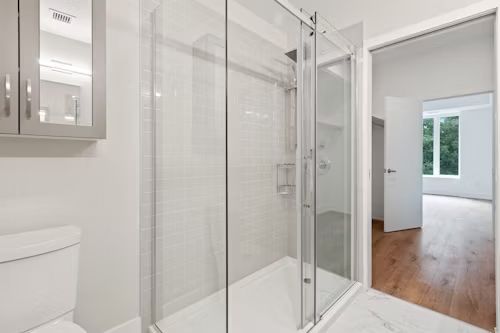 Bathroom with glass shower, toilet, and open door to a room with a window.