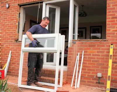 Man installing a white window frame in a brick building's patio. He wears gloves, has a focused expression.