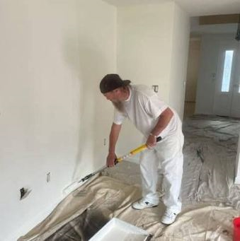 Four workers paint the exterior of a light-gray house white using paint rollers on a wooden deck.