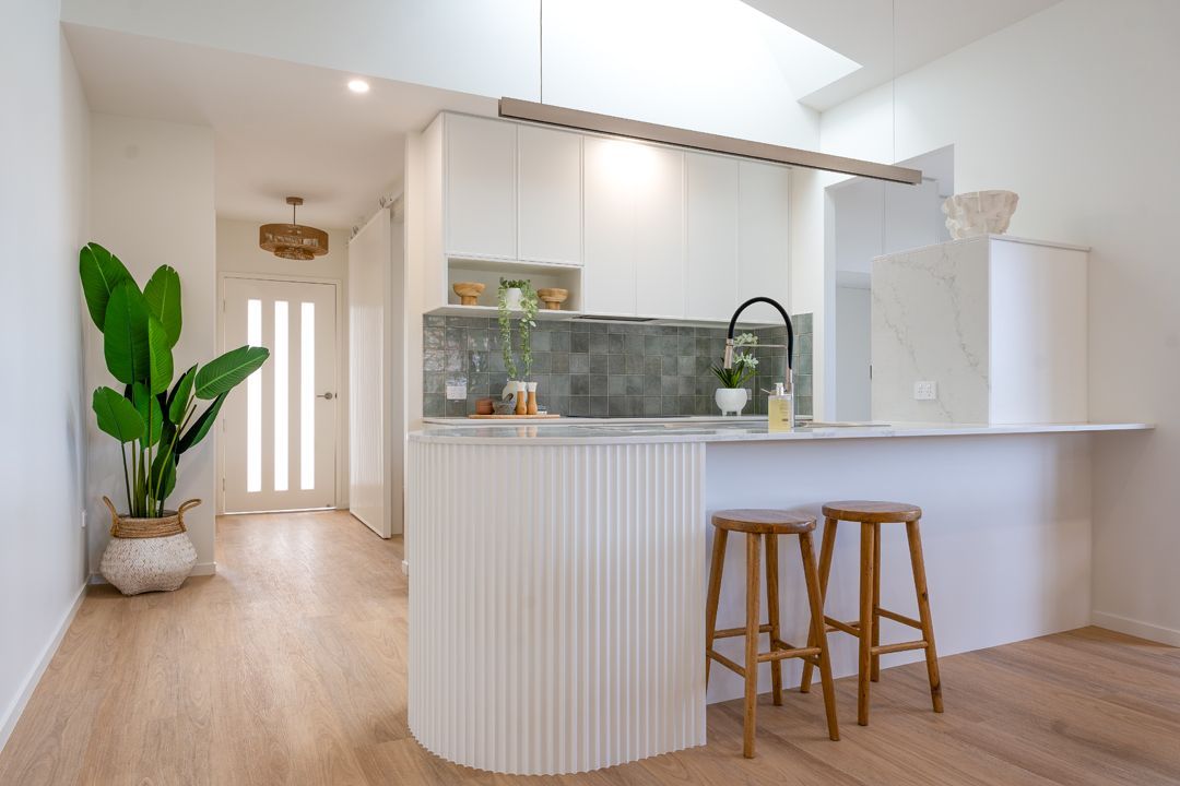 Modern kitchen with a curved counter, bar stools, and a large plant in a bright, open space.