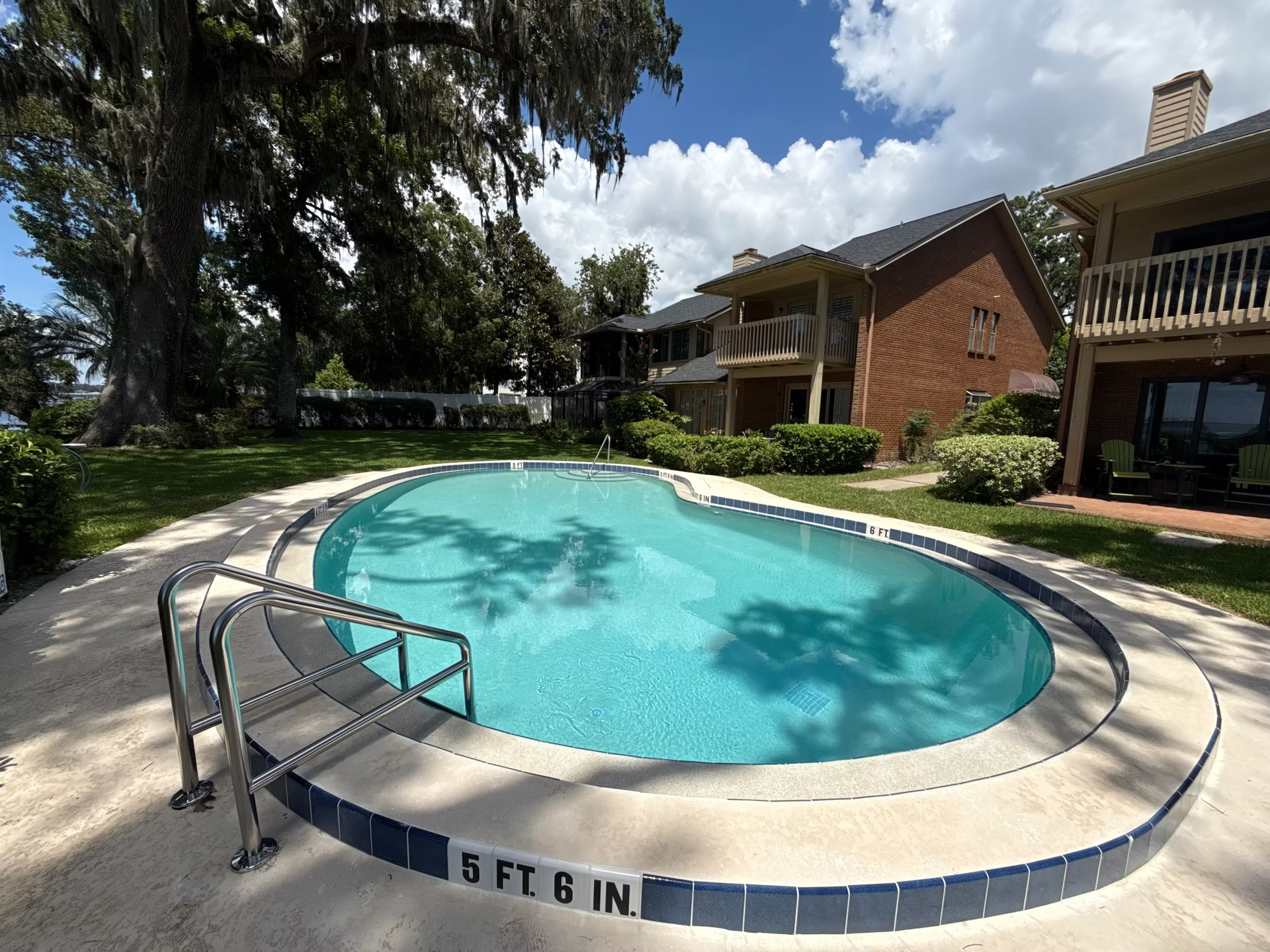 Apartment pool with clear blue water, steps, and lounge chairs beside brick buildings and trees