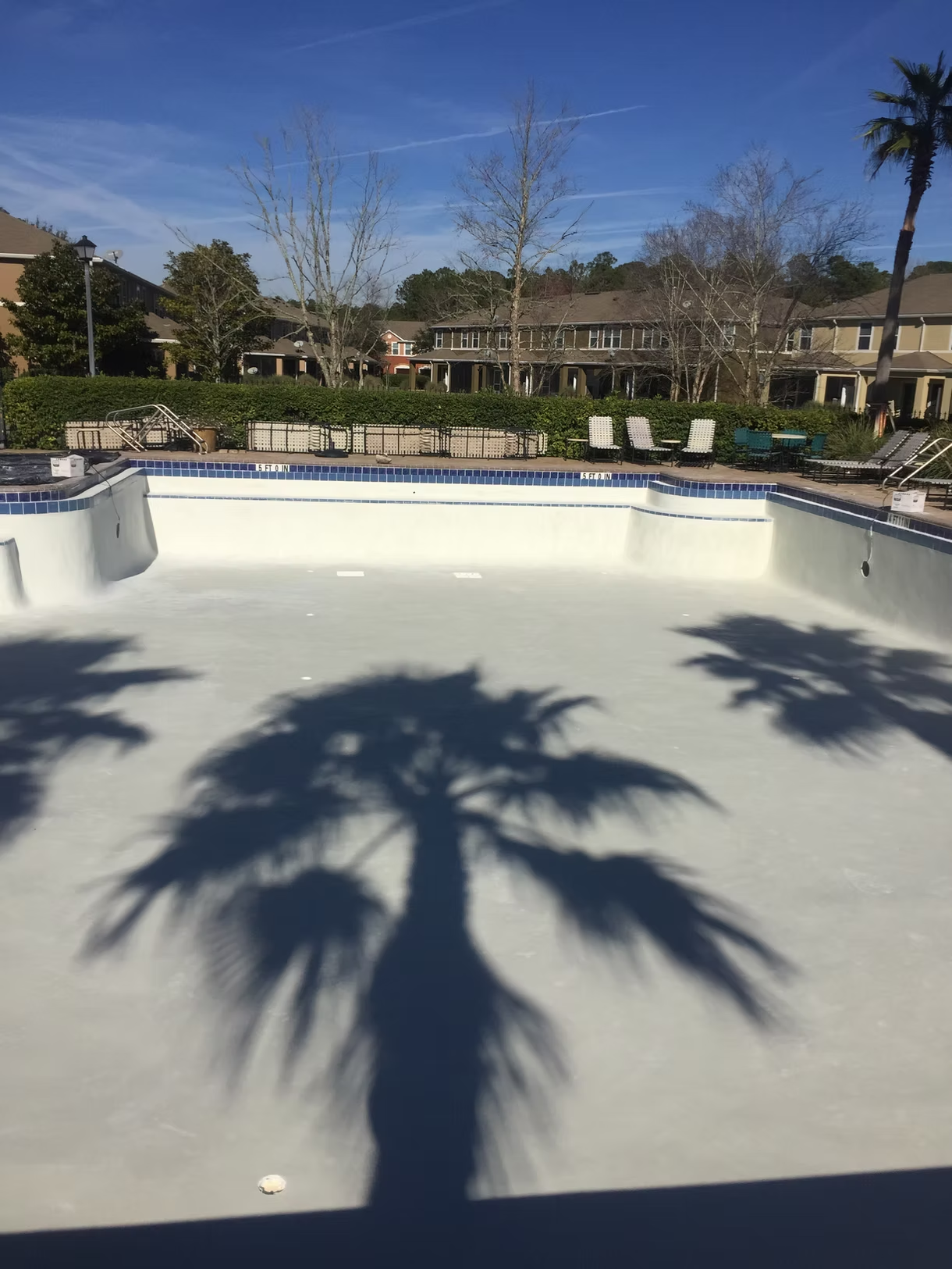 Empty swimming pool with palm tree shadows, blue sky, and lounge chairs in a courtyard.