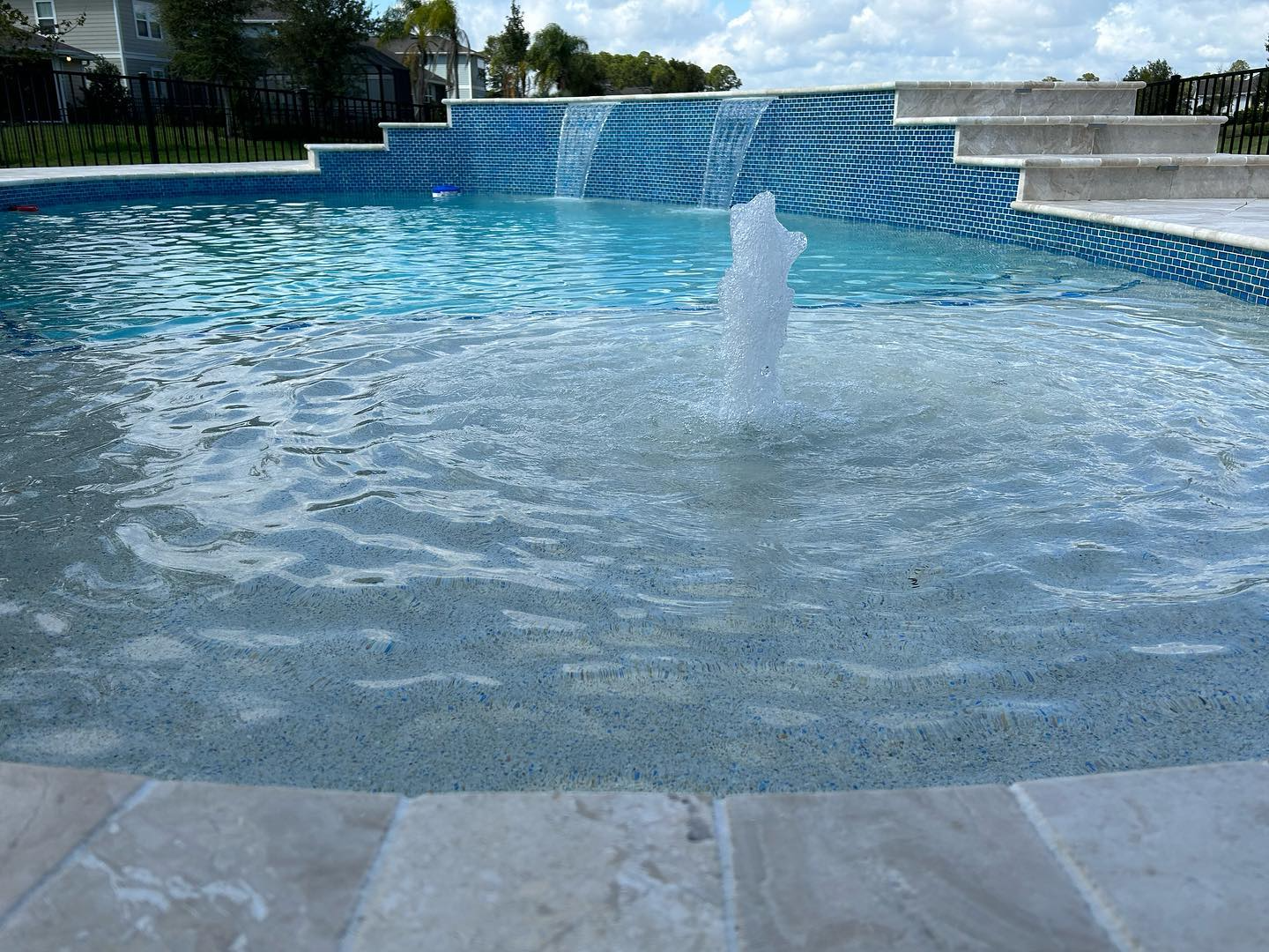 Blue-tiled pool with a central water fountain and stone coping under a sunny sky