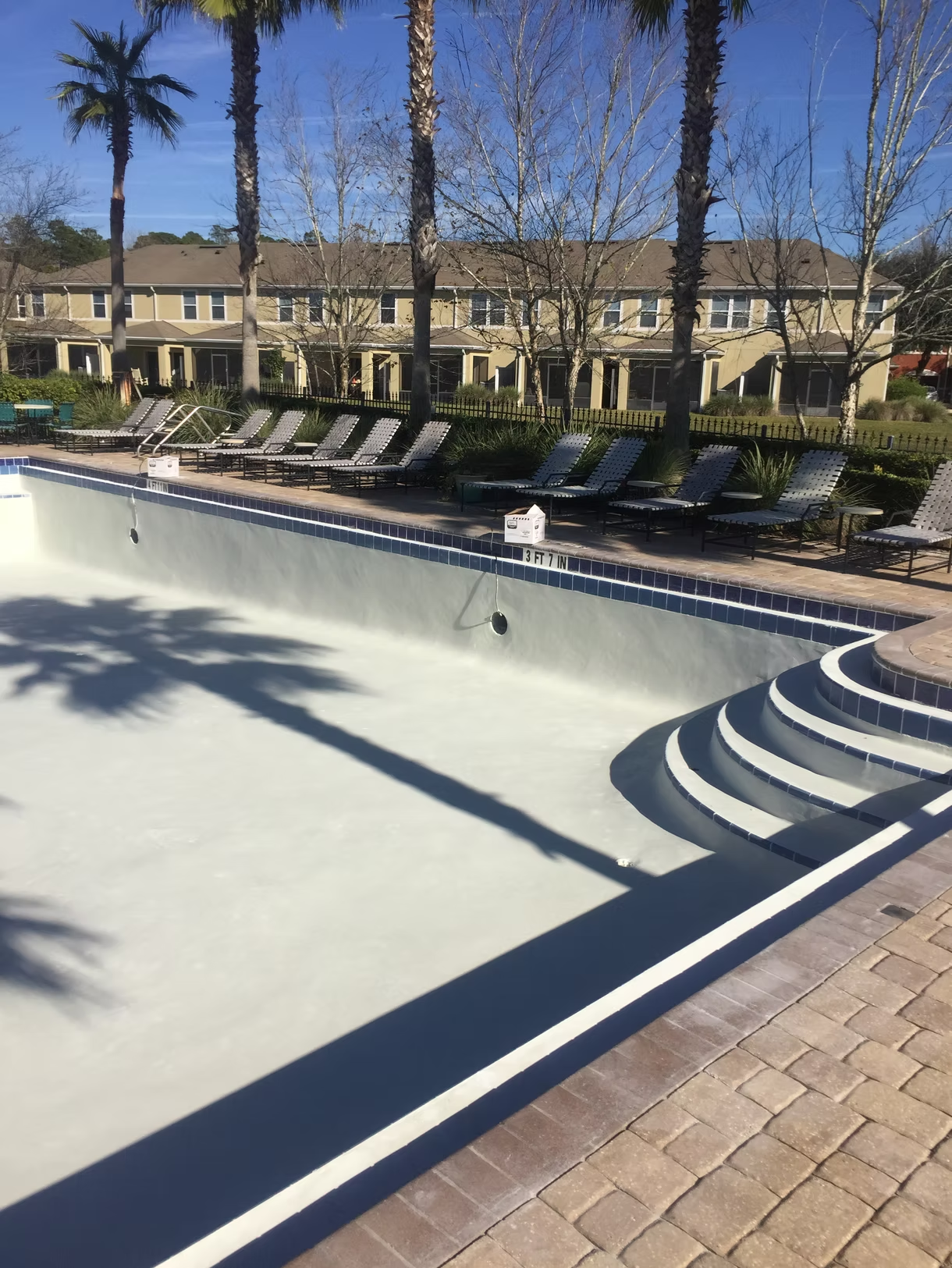 Empty drained swimming pool beside lounge chairs and trees in a sunny courtyard