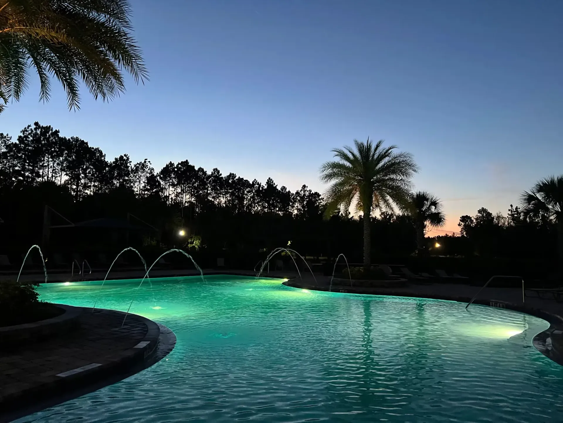 Twilight pool lit turquoise beside palm trees and fountain jets against a darkening sky