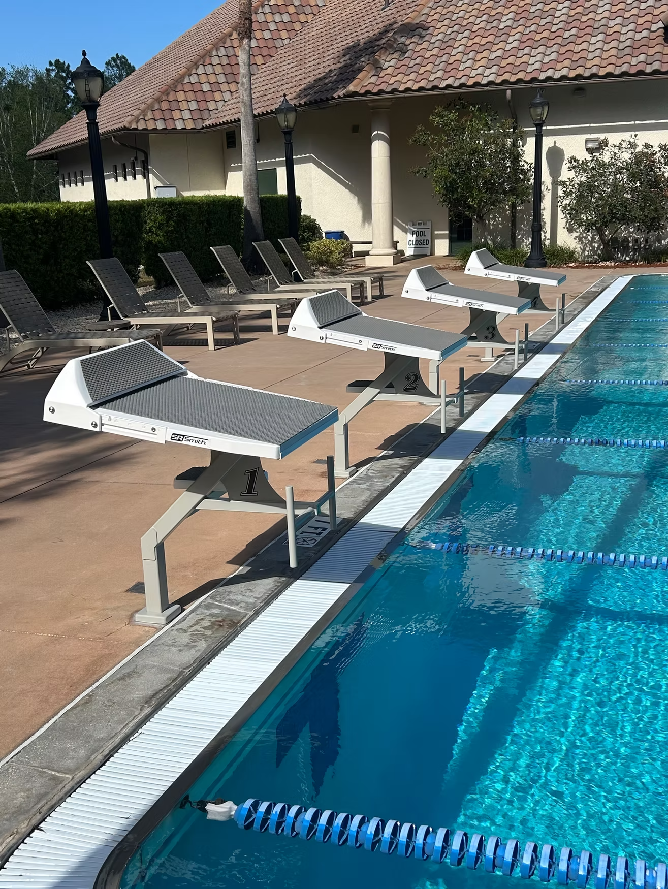 Poolside lounge chairs beside a blue swimming pool at a resort, with umbrellas and a tiled walkway.