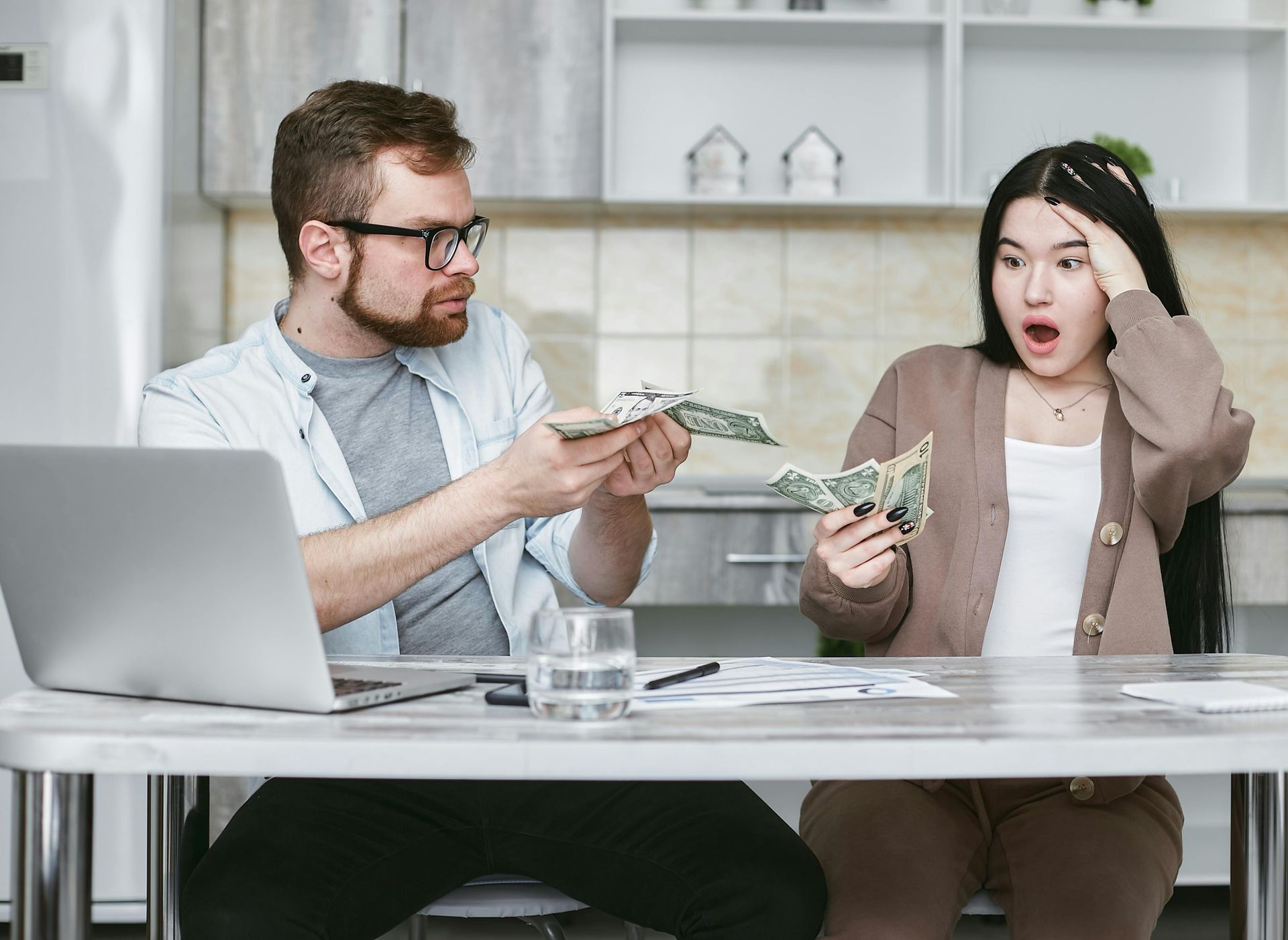 Two people exchanging cash at a desk with a laptop and papers, one looking shocked, the other smiling.