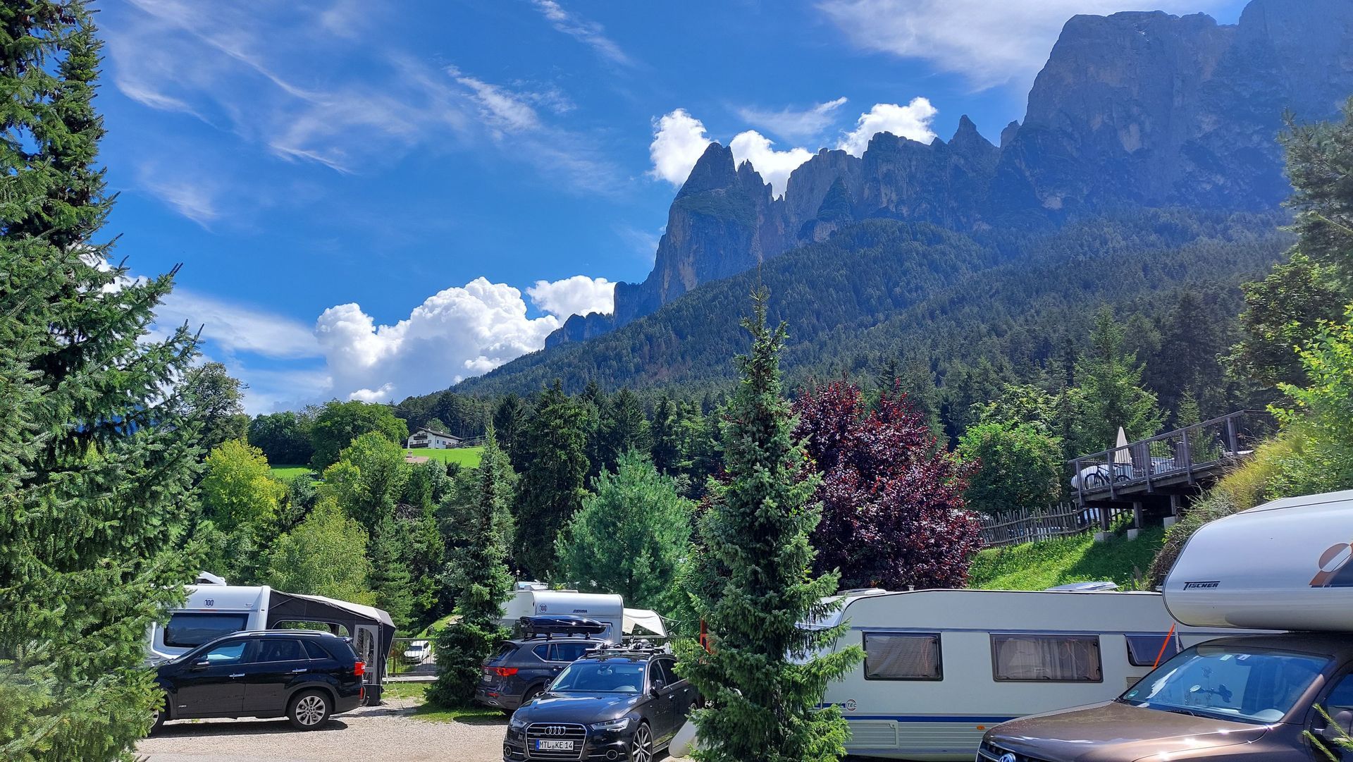 Camping met campers en auto's onder een bergketen, bomen en een blauwe lucht met wolken.