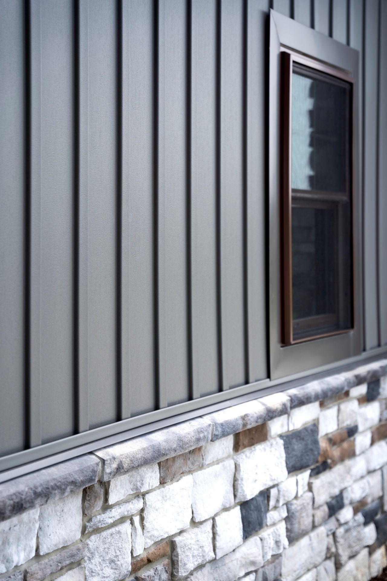 Gray vertical siding, stone foundation, and brown-framed window.