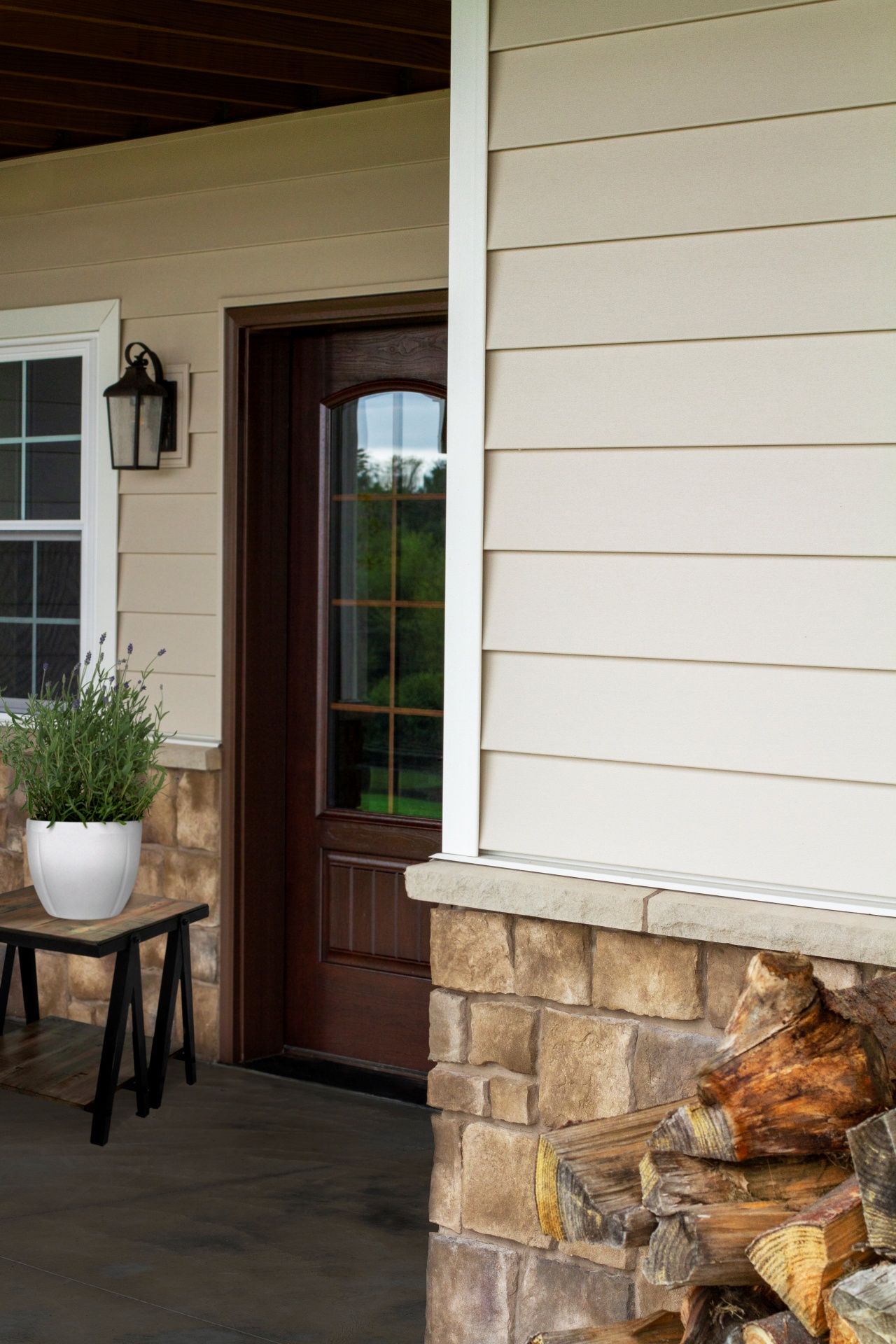 Exterior view of a house porch with a wooden door, beige siding, stone accents, and a potted plant.