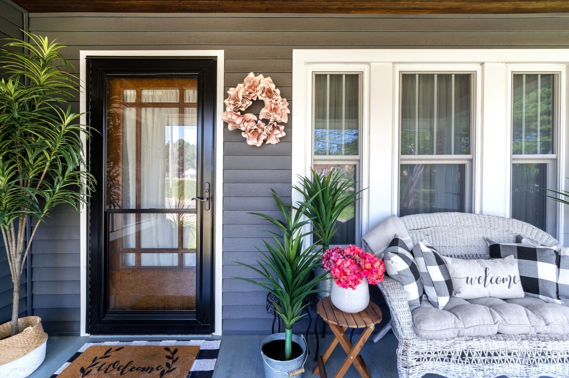 Cozy front porch with black door, pink flowers, wicker furniture, and welcome mat.