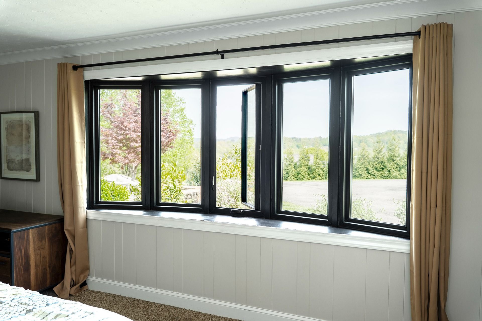Bay window with black frames, tan curtains, and white paneling. Opens to a view of trees and greenery.