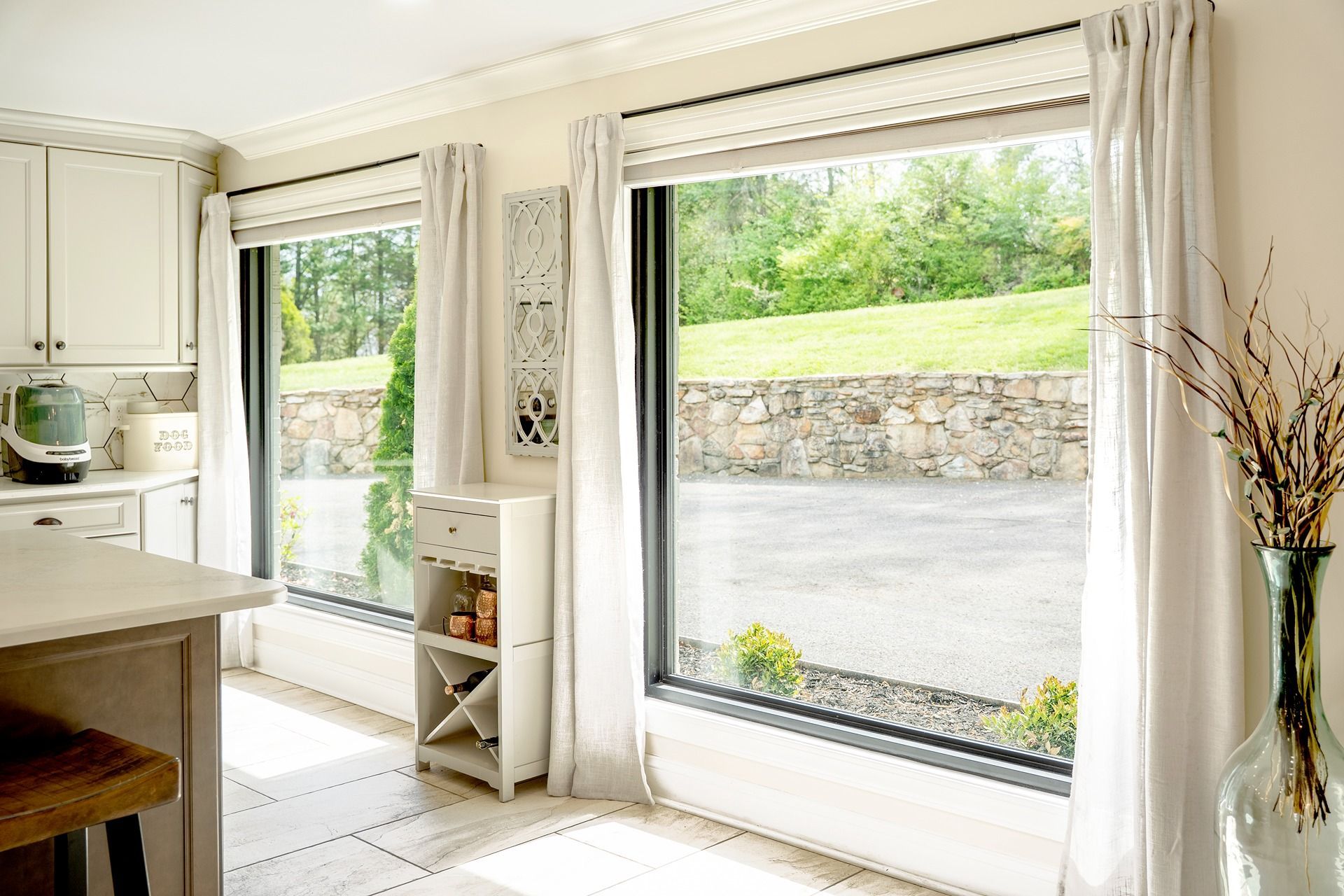 Kitchen with large windows, white cabinetry, and light curtains. Outside, a green yard and stone wall are visible.