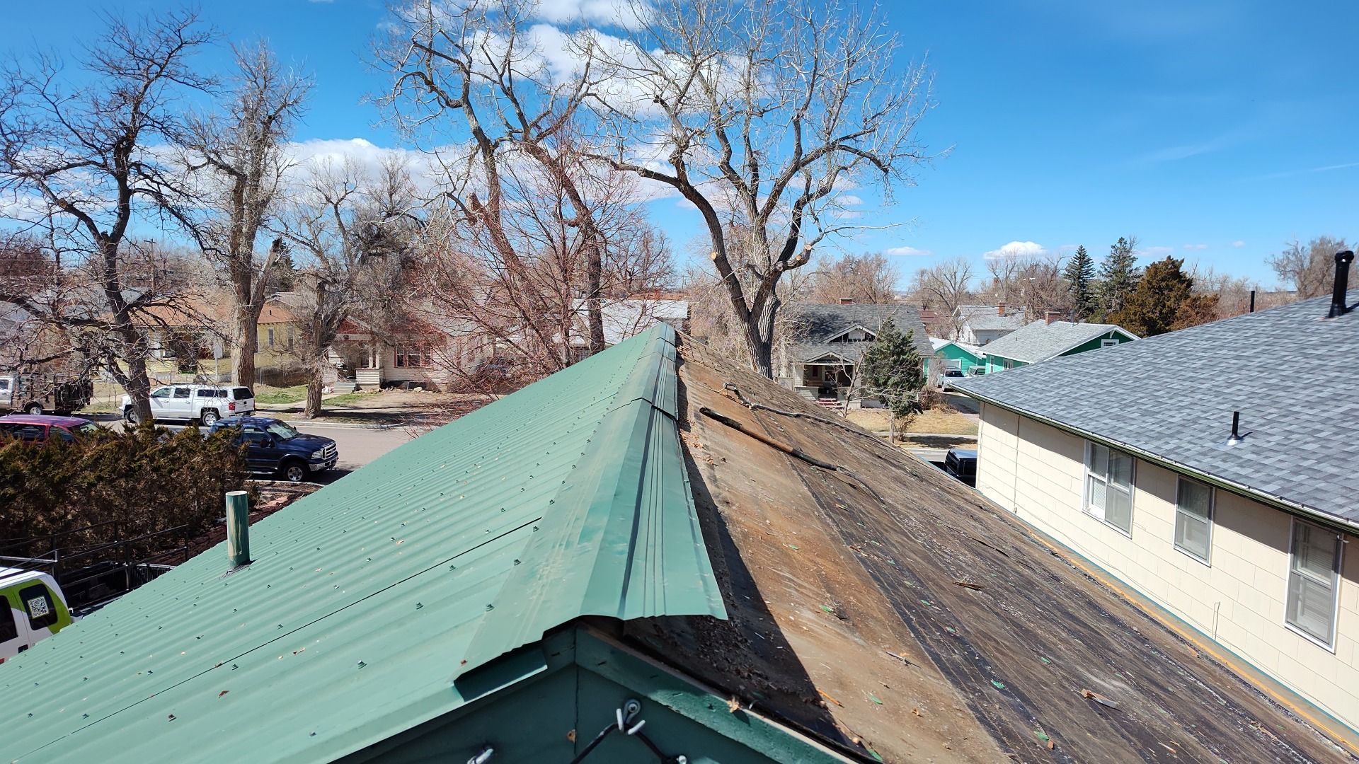 Green metal roof next to a worn, brown roof with trees and houses in the background on a sunny day.