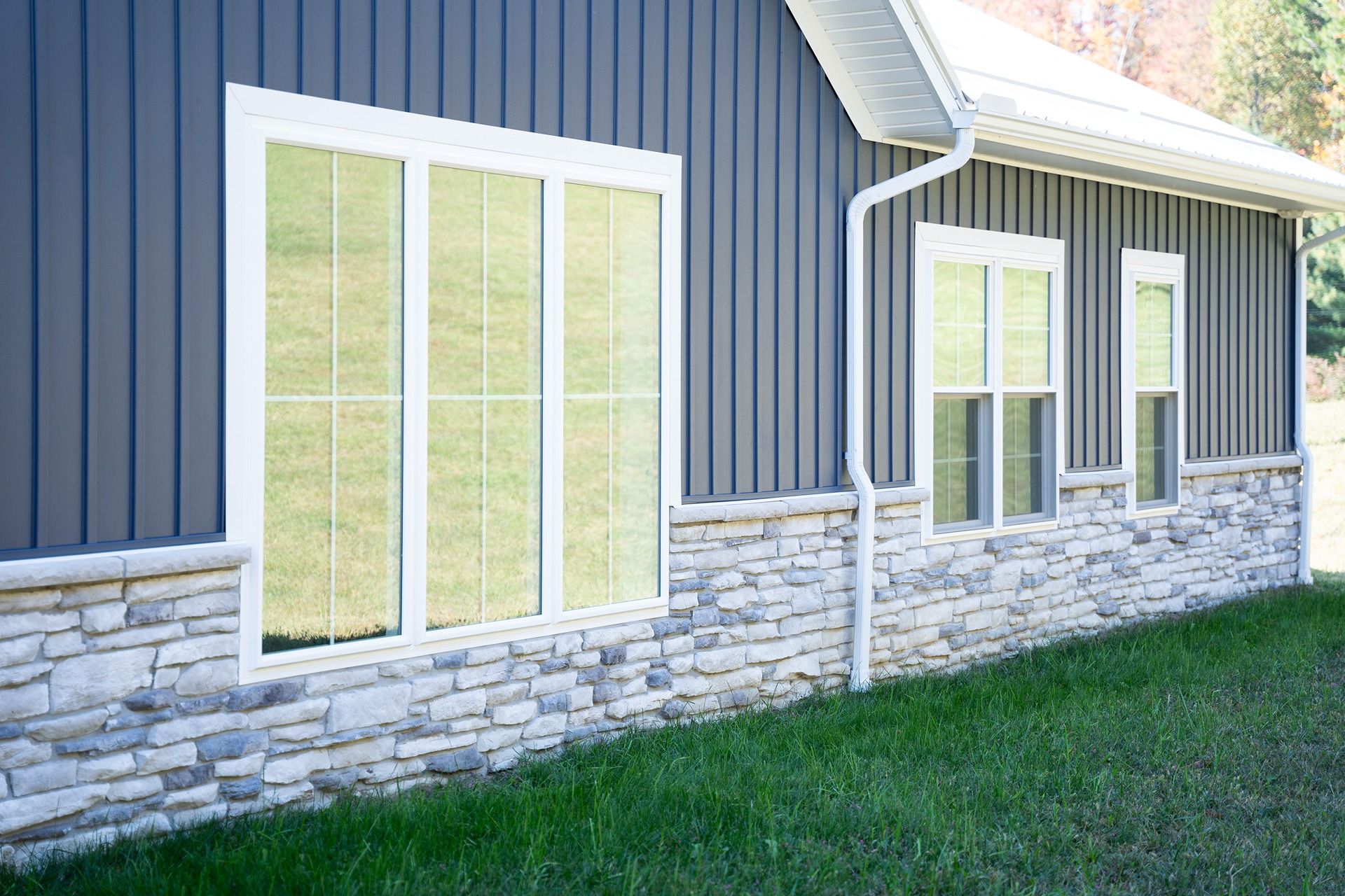 Gray house exterior with stone base, vertical siding, white windows, and green grass.