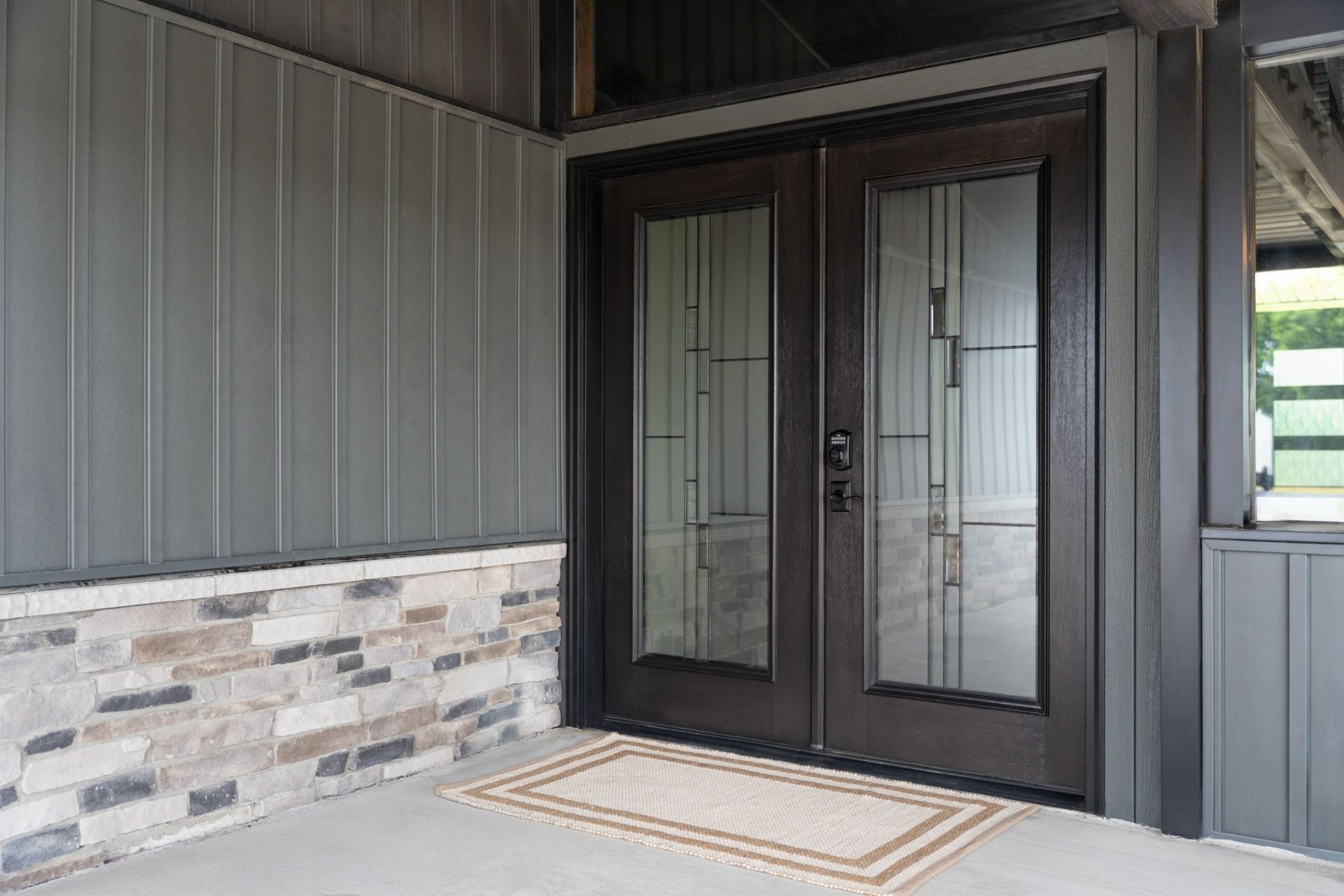 Dark brown double doors with glass panels, on a porch with stone and gray siding.