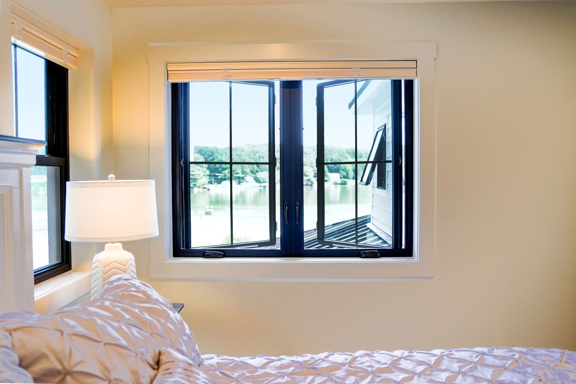 Bedroom interior with a large, black-framed window overlooking a green outdoor view and a lit bedside lamp.