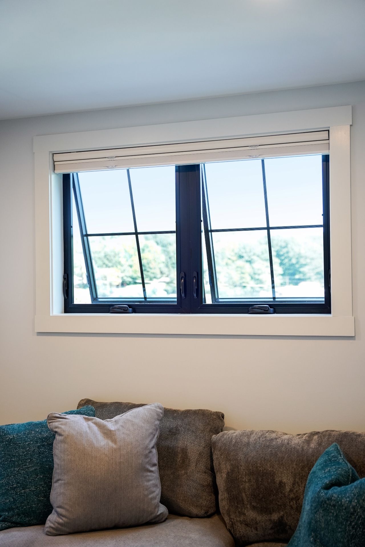 Window with black frame and grid, open, above a sofa with pillows, overlooking a lake.