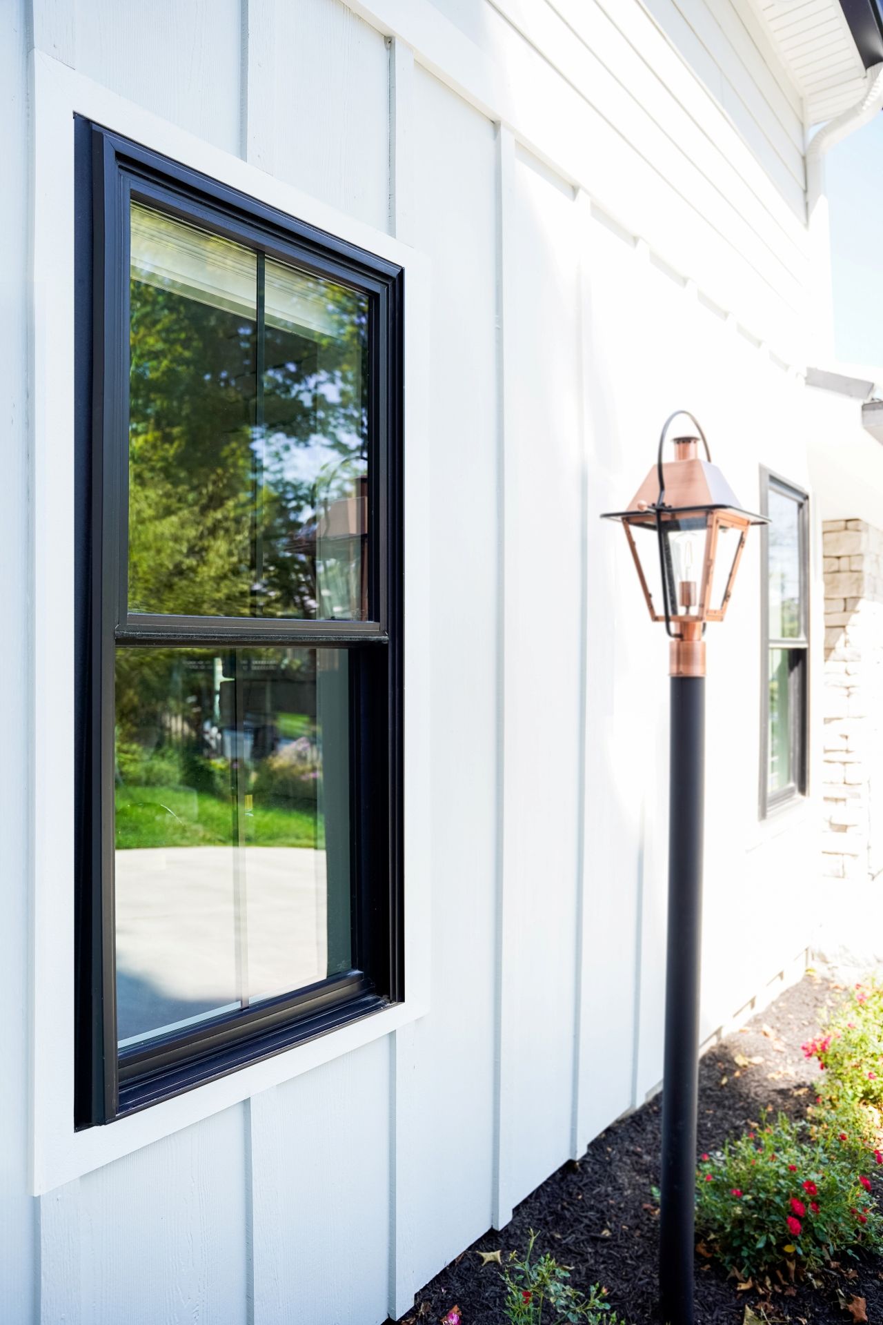 Black-framed window on a white-paneled building with a copper-topped lamp post and bushes.