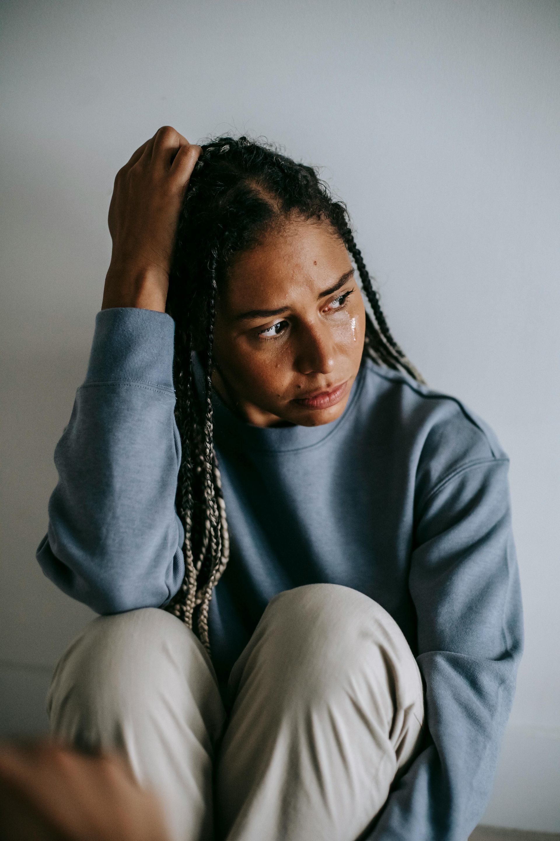 Person with long braids, sitting and holding head, looking distressed. Pale blue sweater, beige pants, white wall.