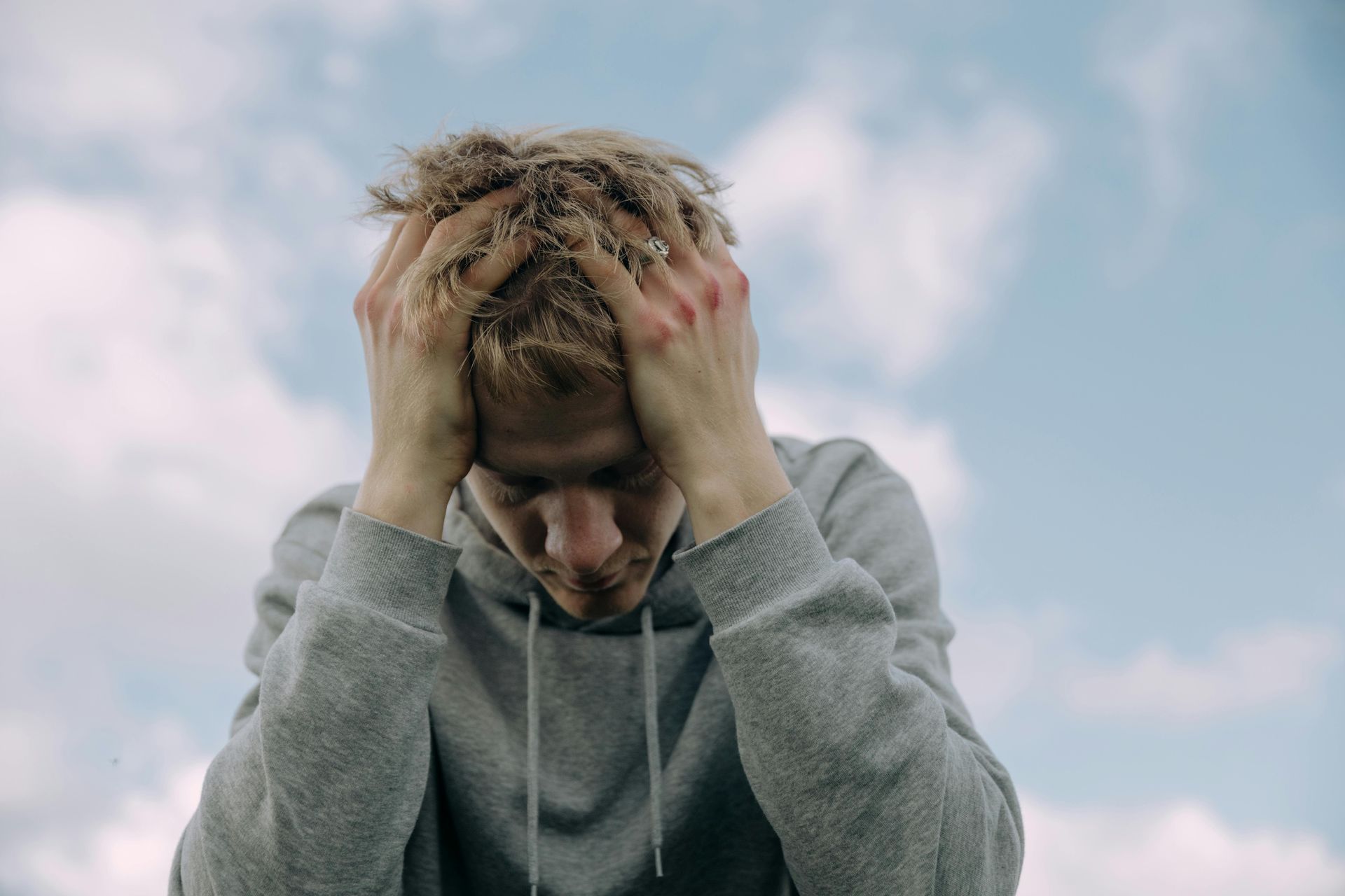 Person in gray hoodie holding head, looking down with a stressed expression; sky background.