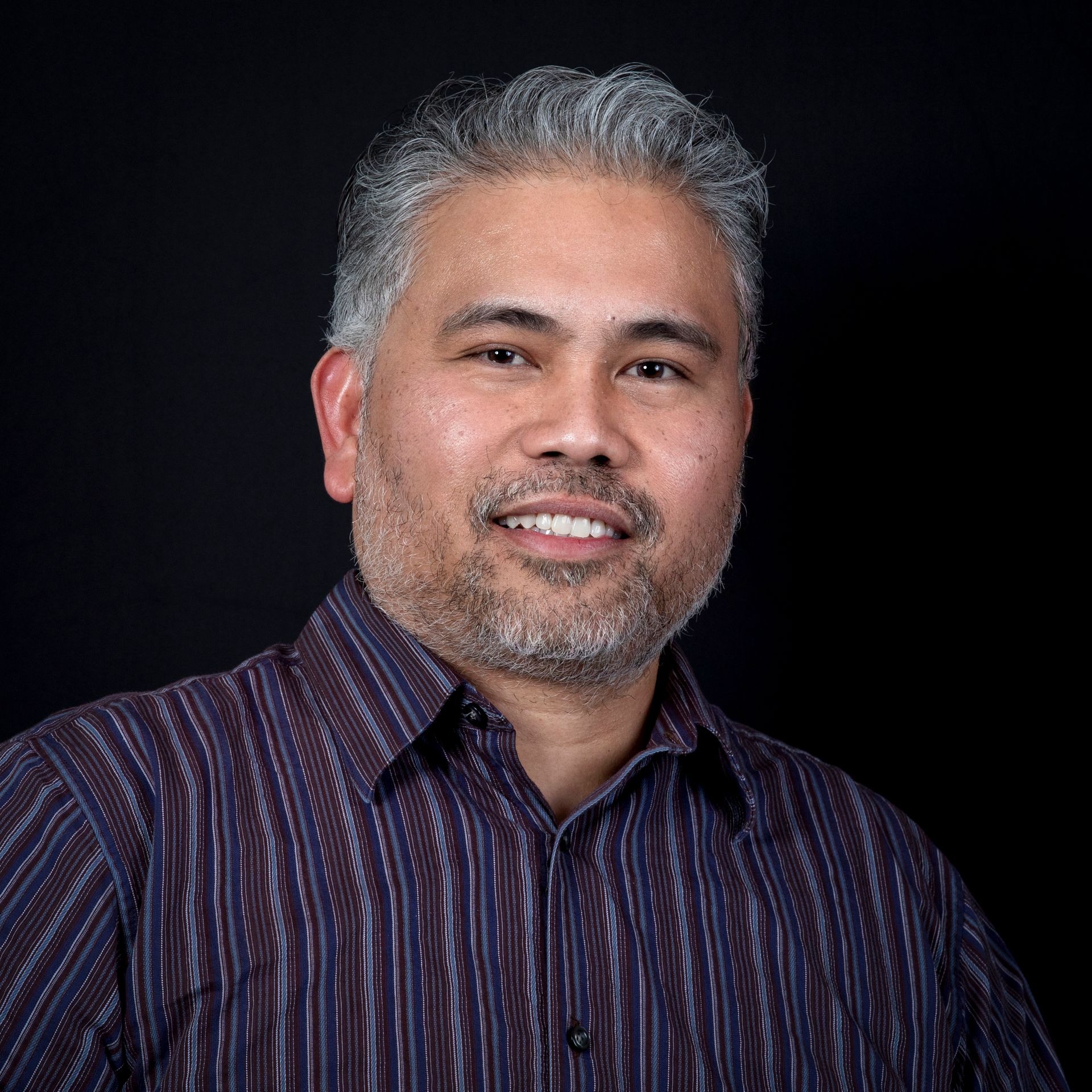 A portrait of a smiling man with short gray hair and a striped shirt on a black background.