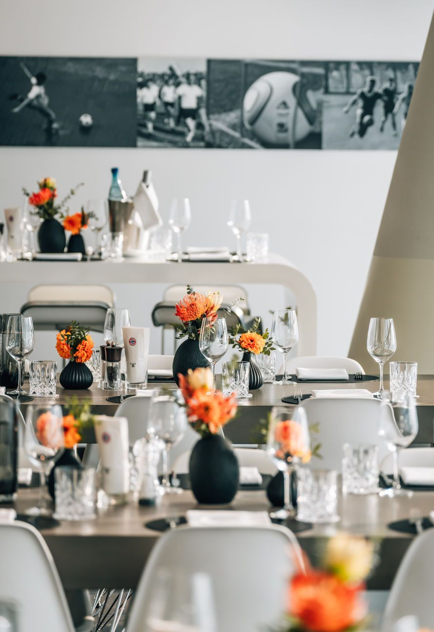Elegante Essgruppe mit weißen Stühlen, orangefarbenen Blumenarrangements, Gläsern und schwarz-weißer Wandkunst im Hintergrund