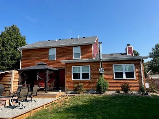 Two-story house with cedar siding, red trim, and gazebo in backyard under a clear blue sky.