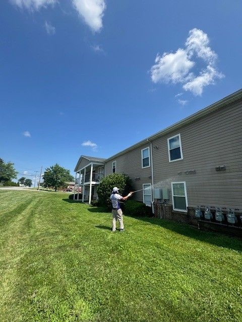 Person spraying siding of a building on a sunny day. Green lawn, blue sky, white clouds.