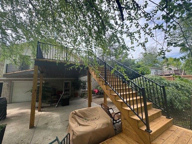Outdoor wooden deck with stairs and black railing, viewed from below, trees in background.