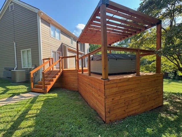 Wooden deck with a hot tub and pergola, next to a house with beige siding and green grass.